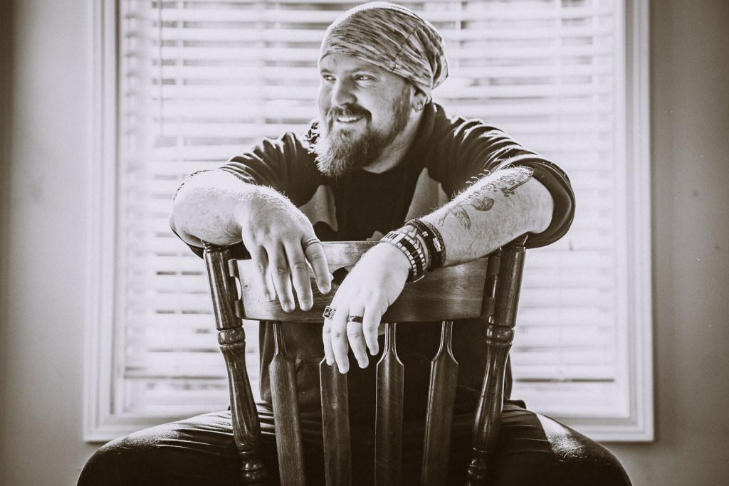 A B&W self portrait of photographer Adam Scott. He sits in a chair backwards, with a beanie on his head smiling.